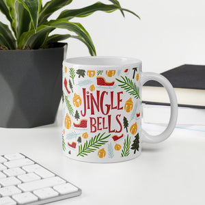 A mug featured on a desk with a plant and a keyboard. The white mug has the words "Jingle Bells" in red lettering and cute Christmas illustrations around the words. The illustrations are sleighs, pine trees, leaves, and ornaments. The illustrations are in the colors light and dark green, light blue and yellow. 