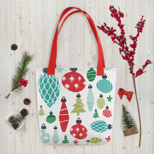 A white tote bag with red handles laying on a table with Christmas items around it. The tote bag features illustrated vintage Christmas ornaments in the colors blue, green and red in various shades. 