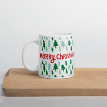 Load image into Gallery viewer, A white mug sitting on a light wood cutting board. The white mug shows the red words &quot;Merry Christmas&quot; in the middle in red letters. The words are surrounded by light and dark green illustrated pine trees. 