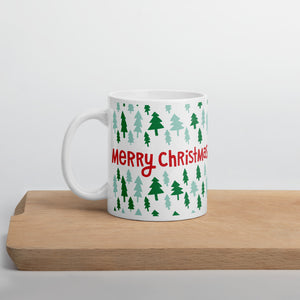 A white mug sitting on a light wood cutting board. The white mug shows the red words "Merry Christmas" in the middle in red letters. The words are surrounded by light and dark green illustrated pine trees. 