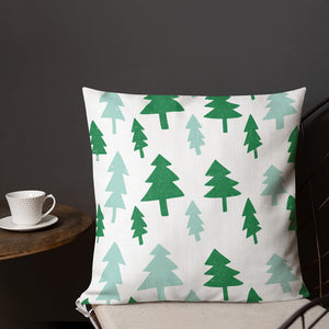 A pillow on a chair with a coffee mug on a table next to it. The white pillow features large illustrated pine trees in the colors dark and light green. 