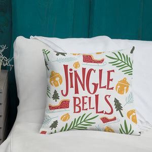 A white pillow with illustrations leading on white bedding with a side table off to the side. The pillow features the words Jingle Bells in red with an illustrated Christmas pattern around the words. The illustrations are a sleigh, leaves, pine trees, and ornaments in the colors yellow, light blue, red, and light and dark green.