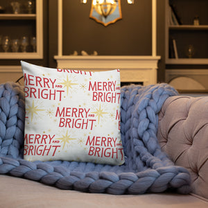 A white pillow on a sofa with a blue knitted blanket. The white pillow features a pattern of the words Merry and Bright with a pattern of Christmas stars. The words in the pattern are red and the stars are yellow. 