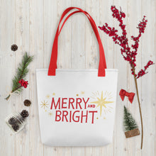 Load image into Gallery viewer, A white tote bag with red handles laying on a table with Christmas items around it. The tote bag features yellow illustrated stars and the words Merry and Bright in red. The words and stars create a pattern all over the tote bag. 