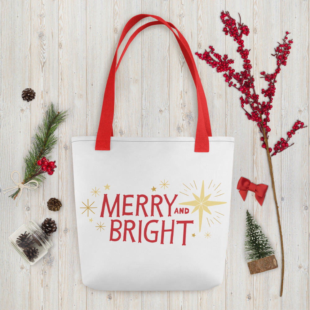 A white tote bag with red handles laying on a table with Christmas items around it. The tote bag features yellow illustrated stars and the words Merry and Bright in red. The words and stars create a pattern all over the tote bag. 