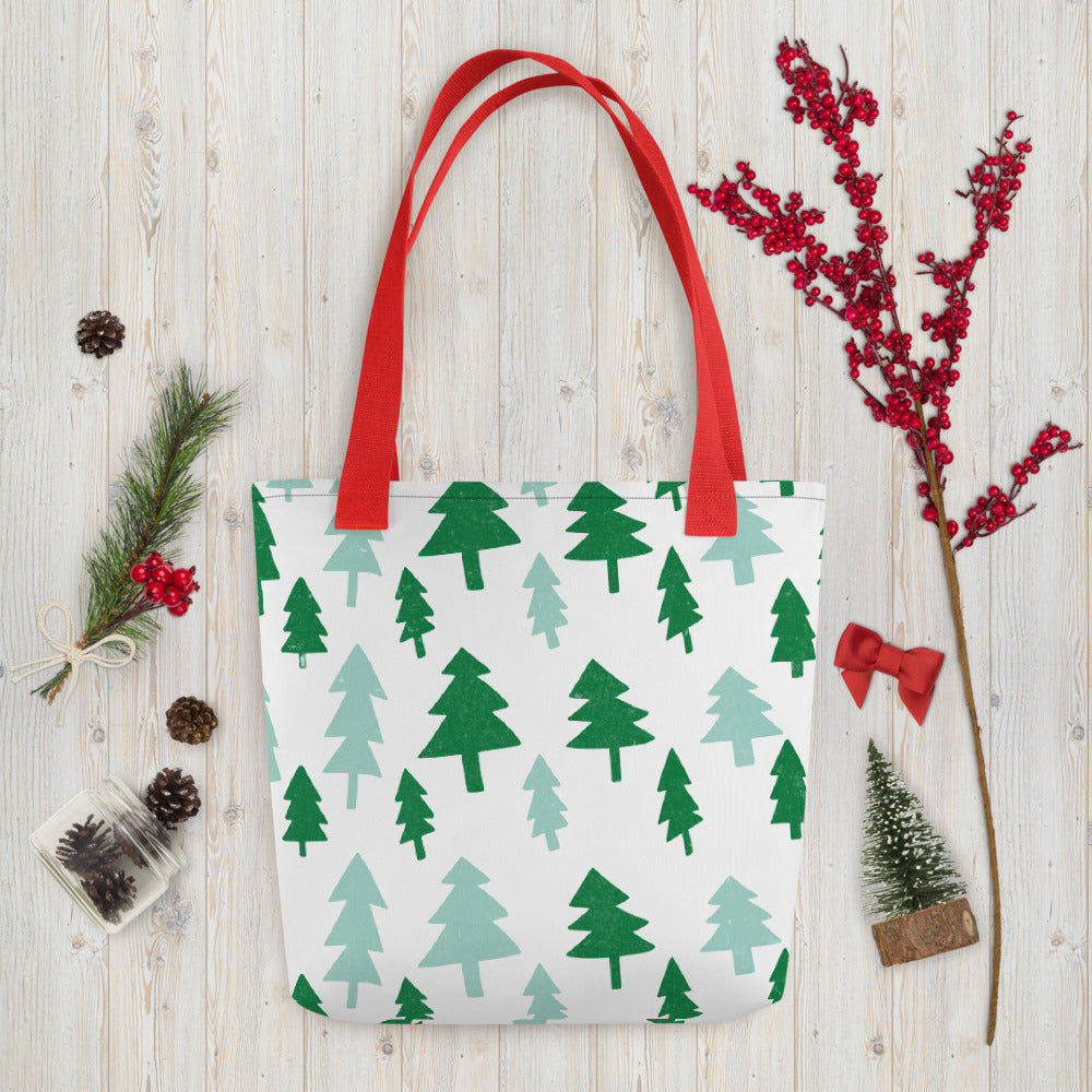 A white tote bag with red handles laying on a table with Christmas items around it. The tote bag features illustrated pine trees in a pattern in dark and light green. 
