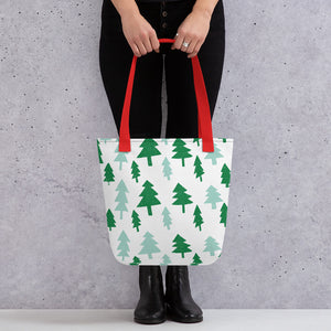 Someone holding a tote bag with red handles and a white fabric bag. The artwork features illustrated Christmas pine trees in light and dark green. 