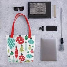 Load image into Gallery viewer, A tote bag lying on a surface with a laptop and office items next to it. The Christmas tote bag features hand illustrated ornaments in an all-over pattern. The pattern colors are in red, blue and green. 