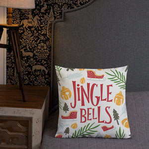 A pillow leaning on a grey headboard with a table and lamp off to the side. The white pillow features Christmas illustrations with the words Jingle Bells in the middle in red. The illustrations featured are ornaments, sleighs, pine trees and leaves. The illustrations are in the colors light and dark green, light blue, yellow and red. 