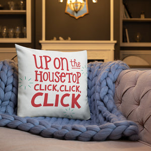 A white pillow on a sofa with a blue knitted blanket. The white pillow features the words "Up on the housetop, click, click, click." The words are in red with three blue star patterns around the words. 