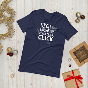 A navy T-shirt laying on the ground with Christmas items surrounding it. The T-shirt features the words  "Up on the housetop, click, click, click" in white. There are three stars around the words in white. 