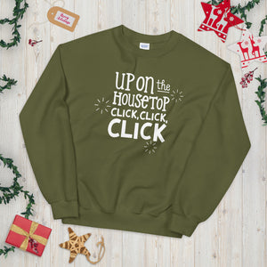 An olive green sweatshirt laying on a table with Christmas objects around it. The sweatshirt features the words "Up on the housetop, click, click, click" in white. There are three white stars around the letters. 