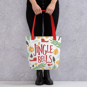 Someone holding a tote bag with red handles and a white fabric bag. The artwork features illustrated Christmas items including sleighs, ornaments, leaves and pine trees. The illustrations are in the colors light and dark green, light blue, yellow and red. The words read Jingle Bells in red. 
