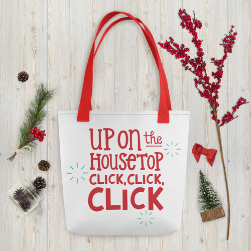 A white tote bag with red handles laying on a table with Christmas items around it. The tote bag features the words 