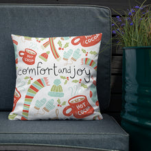 Load image into Gallery viewer, The white pillow is leaning on a sofa with a plant off to the side. The pillow is white with Christmas illustrations featuring mittens, hot cocoa mugs, holy leaves, winter hats and socks. The words Comfort and Joy are in the center of the pillow in black. The  illustrations are in light green, light blue, and red.