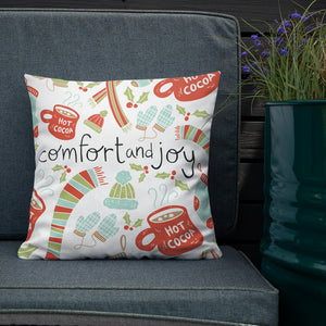 The white pillow is leaning on a sofa with a plant off to the side. The pillow is white with Christmas illustrations featuring mittens, hot cocoa mugs, holy leaves, winter hats and socks. The words Comfort and Joy are in the center of the pillow in black. The  illustrations are in light green, light blue, and red.