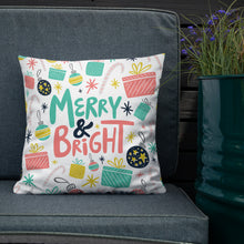 Load image into Gallery viewer, The white pillow is leaning on a sofa with a plant off to the side. The pillow is white with Christmas illustrations featuring gifts, ornaments, candy canes and stars. The words Merry &amp; Bright are in the center of the pillow. The words and illustrations are in pink, yellow, light blue and black. 