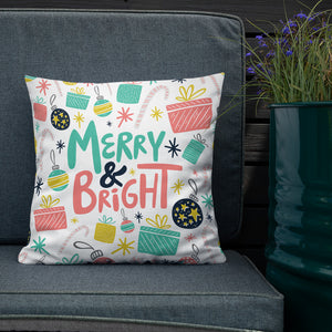The white pillow is leaning on a sofa with a plant off to the side. The pillow is white with Christmas illustrations featuring gifts, ornaments, candy canes and stars. The words Merry & Bright are in the center of the pillow. The words and illustrations are in pink, yellow, light blue and black. 