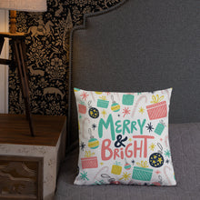 Load image into Gallery viewer, A pillow leaning on a grey headboard with a table and lamp off to the side. The white pillow features Christmas illustrations with candy canes, stars, presents and ornaments. The words Merry &amp; Bright are in the center of the pillow. The colors of the pattern and words are yellow, pink, light blue and black. 