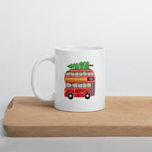 Load image into Gallery viewer, A white mug sitting on a light wood cutting board. The white mug features a hand illstrated double decker London bus with a Christmas tree on top. The is red and the tree is green top. 