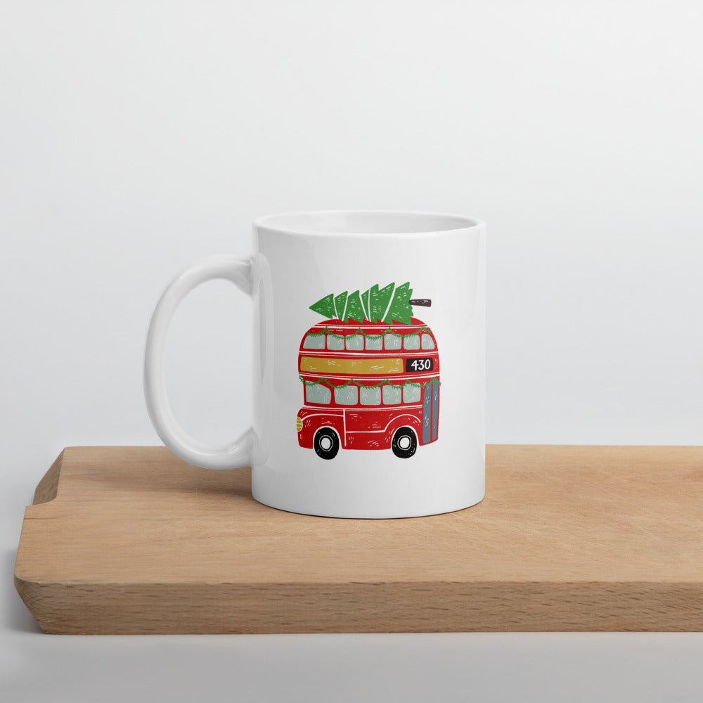 A white mug sitting on a light wood cutting board. The white mug features a hand illstrated double decker London bus with a Christmas tree on top. The is red and the tree is green top. 