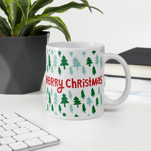 A mug featured on a desk with a plant and a keyboard. The white mug has the words "Merry Christmas" in red lettering and illustrated pine trees around them. The pine trees are in light and dark green. 