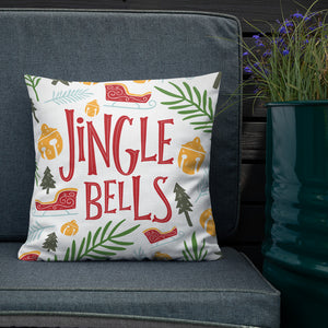 The white pillow is leaning on a sofa with a plant off to the side. The pillow is white with illustrations and wording. The words featured are "Jingle Bells" in the color red with Christmas illustrations around the words. The illustrations are in yellow, light blue, light and dark green and red with sleighs, leaves, pine trees and ornaments. 