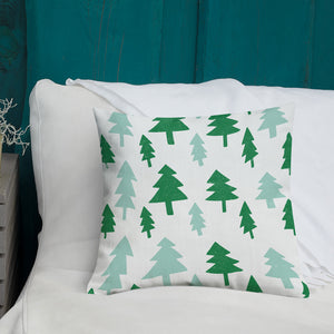 A white pillow with illustrations leading on white bedding with a side table off to the side. The white pillow features illustrated pine trees in dark and light green. 