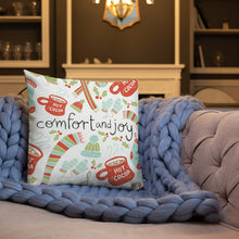 Load image into Gallery viewer, A white pillow on a sofa with a blue knitted blanket. The white pillow features a Christmas pattern. The pattern includes the words Comfort &amp; Joy in the middle in black with an illustrated pattern around the words with hot cocoa mugs, winter hats, mittens, holy leaves and socks. The colors of the patterns are in light blue, light green and red.