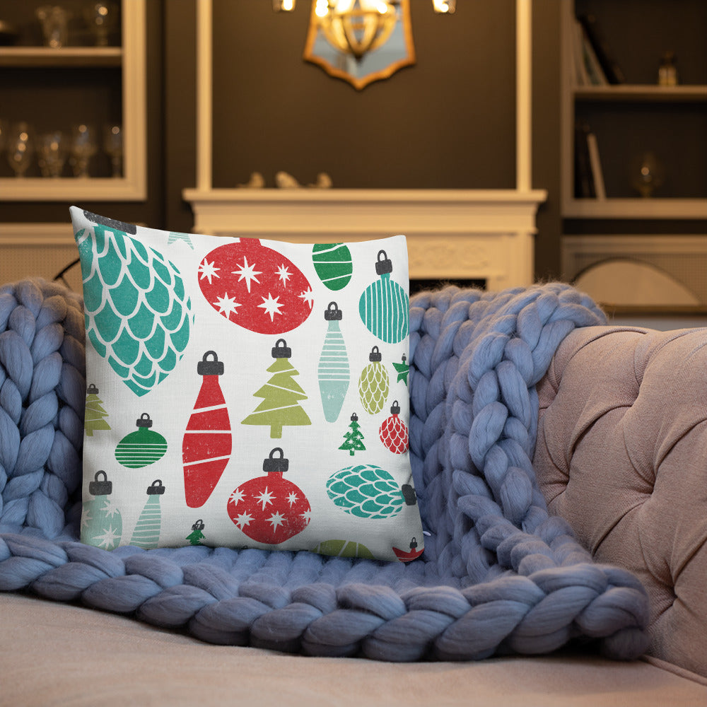 A white pillow on a sofa with a blue knitted blanket. The white pillow features an illustrated ornament pattern featured all over the fabric of the pillow. The colors of the ornaments are red, light green and dark green, blue and light blue. 