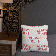 Load image into Gallery viewer, A pillow leaning on a grey headboard with a table and lamp off to the side. The white pillow features a Christmas pattern with the words &quot;Merry and Bright&quot; in red and illustrated stars in red. 
