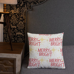 A pillow leaning on a grey headboard with a table and lamp off to the side. The white pillow features a Christmas pattern with the words "Merry and Bright" in red and illustrated stars in red. 