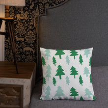 Load image into Gallery viewer, A pillow leaning on a grey headboard with a table and lamp off to the side. The white pillow features Christmas illustrations with pine trees in dark and light green. 