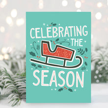 Load image into Gallery viewer, A Christmas card standing up with with pine leaves in the background with a touch of snow.  The Christmas card has a light teal background with a red illustrated sleigh with holly leaves and berries around the sleigh. The words &#39;celebrating the season&#39; are around the sleigh illustrated in white with a touch of red on the inside of the letters. 