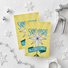 Load image into Gallery viewer, Two Christmas cards laying on a white background with white and silver Christmas decorations on the table. The Christmas card has a yellow background with the words &#39;shine bright this Christmas&#39; in blue and white. There&#39;s an illustrated vintage star Christmas tree topper featured in between the words.
