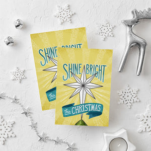 Two Christmas cards laying on a white background with white and silver Christmas decorations on the table. The Christmas card has a yellow background with the words 'shine bright this Christmas' in blue and white. There's an illustrated vintage star Christmas tree topper featured in between the words.