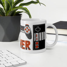 Load image into Gallery viewer, A mug featured on a desk with a plant and a keyboard. The white mug features hand illustrated images of game controllers and the word gamer in black, red and grey. 