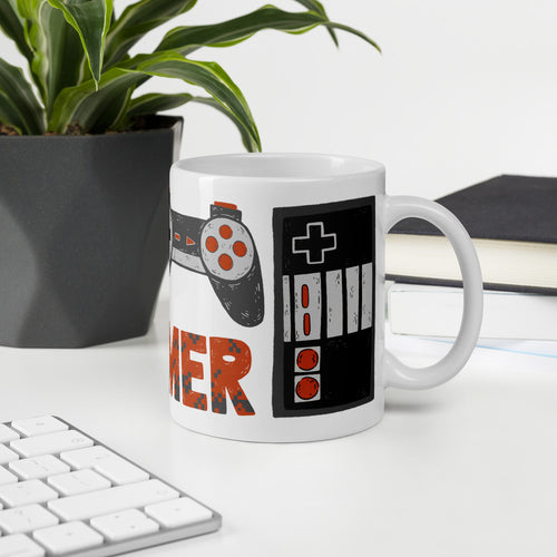 A mug featured on a desk with a plant and a keyboard. The white mug features hand illustrated images of game controllers and the word gamer in black, red and grey. 