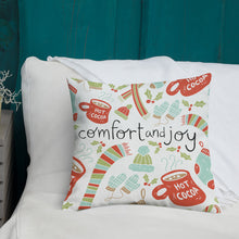 Load image into Gallery viewer, A white pillow with illustrations leading on white bedding with a side table off to the side. The white pillow features illustrated gloves, socks, hot cocoa mugs, winter hats and holy leaves. The middle of the pillow has the words Comfort &amp; Joy with the Christmas pattern around it. The colors of the pattern are light blue, red, and light green. The words are in black. 