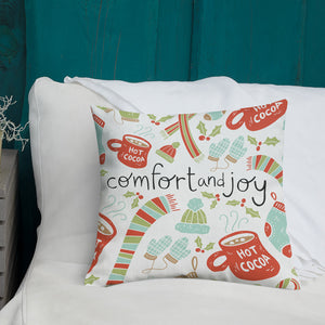 A white pillow with illustrations leading on white bedding with a side table off to the side. The white pillow features illustrated gloves, socks, hot cocoa mugs, winter hats and holy leaves. The middle of the pillow has the words Comfort & Joy with the Christmas pattern around it. The colors of the pattern are light blue, red, and light green. The words are in black. 