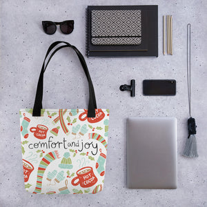 A tote bag lying on a surface with a laptop and office items next to it. The patterned tote bag has a black handle and the tote is white. The pattern includes the words Comfort & Joy in black in the middle with an illustrated pattern around the words with winter hats and gloves, scarves, hot cocoa mugs and holy leaves. The colors of the pattern are in red, light green and light blue. 
