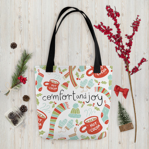 A white tote bag with black handles laying on a table with Christmas items around it. The tote bag features illustrated scarves, winter hats, mittens, hot cocoa mugs and holy leaves. The words Comfort & Joy are in the center of the pattern. The colors of the illustrated patterns are in light blue, light green and red. The words are featured in black. 