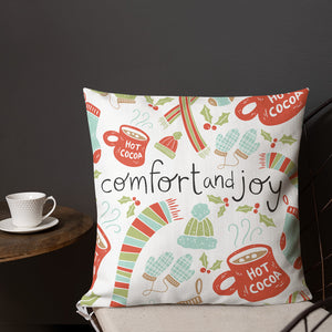 A pillow on a chair with a coffee mug on a table next to it. The white pillow features Christmas illustrations of hot cocoa mugs, scarves, winter hats and gloves, socks, and holy leaves. The illustrations are in light blue, light green, and red. The words Comfort & Joy are in the middle in black. 