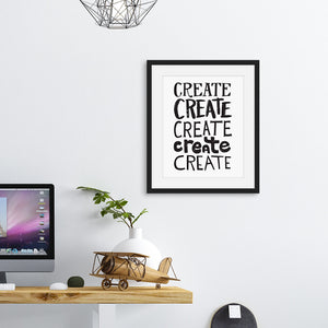 Artwork featured in a black frame above a desk. The artwork is on a white paper with black lettering in the word “create” written five times. 