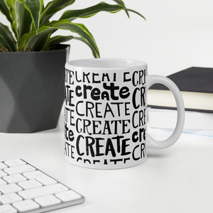 A mug featured on a desk with a plant and a keyboard. The white mug features the phrase “create" repeated over and over in different hand lettered fonts. 