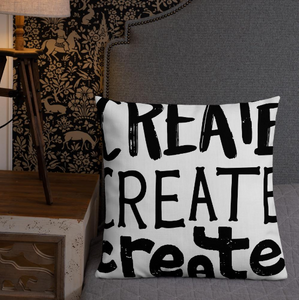A pillow leaning on a grey bed headboard. The white pillow features the phrase “create, create, create" in black lettering with each word in three different fonts. 