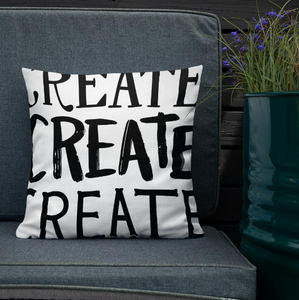 A pillow leaning on a grey sofa with a plant in the background. The white pillow features the phrase “create create create” in black letters.