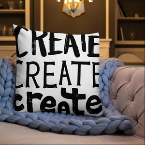 A pillow on a sofa with a blue knitted blanket with the phrase “create, create, create" in black lettering with each word in three different fonts. 