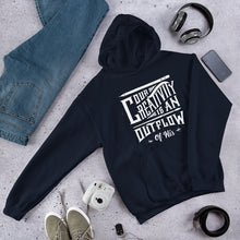 Load image into Gallery viewer, A navy hoodie laying on the ground with objects around it. The hoodie features hand drawn lettering in white with the words &quot;Our creativity is an outflow of His.&quot;