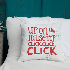 A white pillow with illustrations leading on white bedding with a side table off to the side. The white pillow has the words "Up on the housetop, click, click, click" in red lettering. There are three star patterns in blue around the words. 